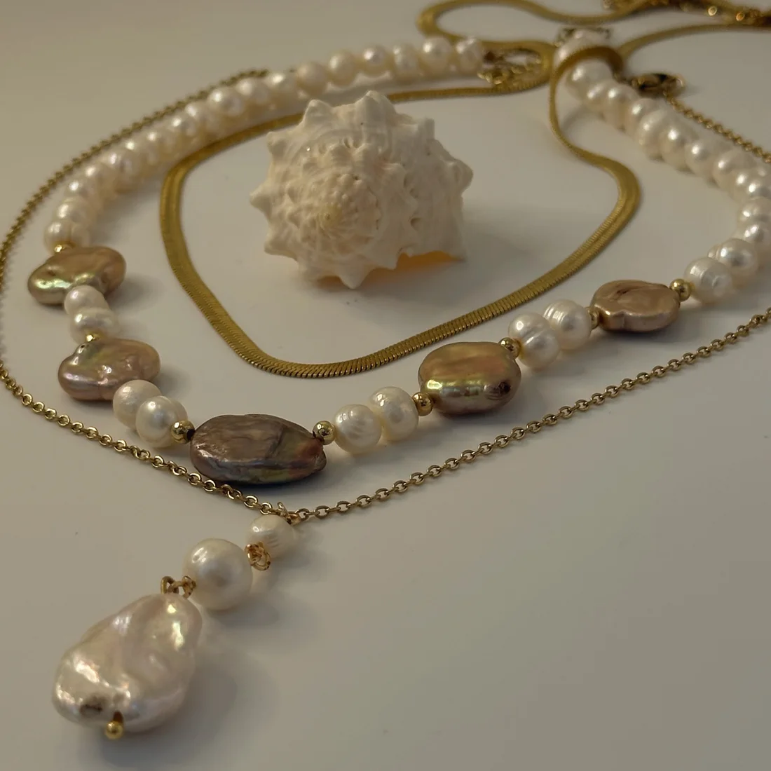 Collares de perlas barrocas y cadena dorada junto a un caracol.